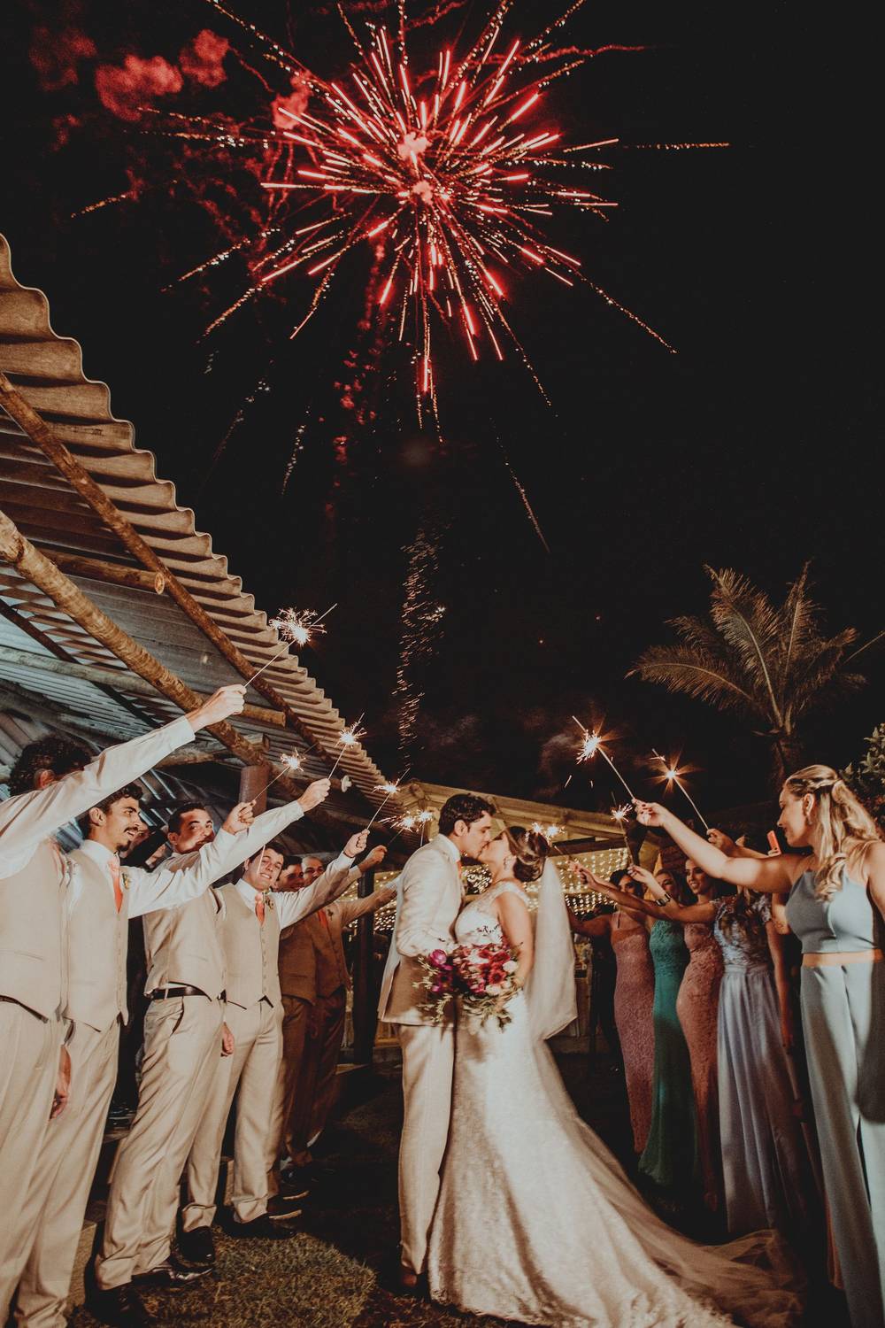 A bride and groom kissing under red fireworks while their guests hold up sparklers